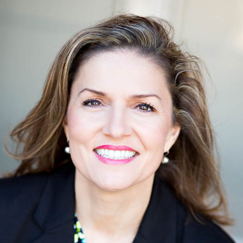 A woman with long brown hair and pearl earrings is smiling at the camera, wearing a black blazer over a patterned top, with a neutral background.