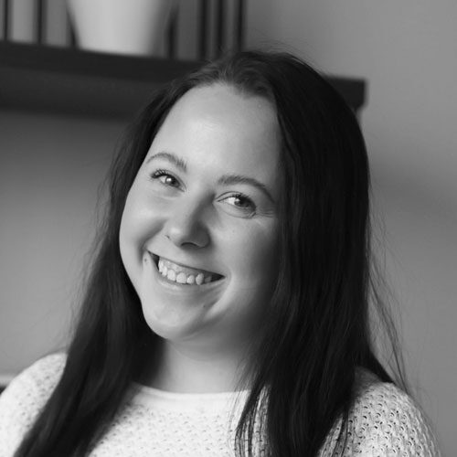 A woman with long dark hair smiles at the camera. She is wearing a light textured top, and there is a shelf with a plant in the background.